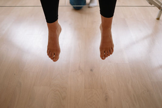 Bare feet with unpainted natural toenails hanging above a pale wooden floor indoors